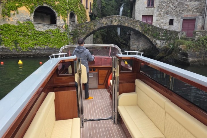 Person on a boat approaching a stone bridge by ivy-covered buildings and a waterfall.