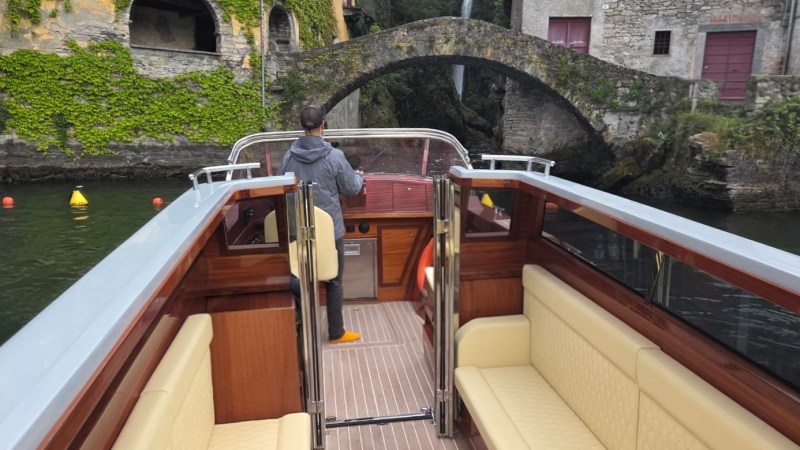 Person on a boat approaching a stone bridge by ivy-covered buildings and a waterfall.