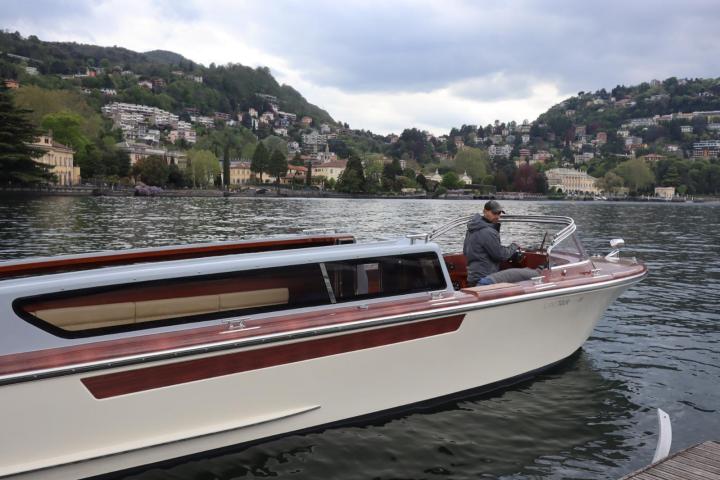 Person steering a boat on a lake with hills and houses in the background.