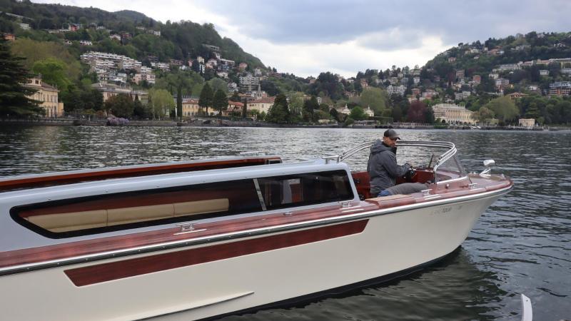 Person steering a boat on a lake with hills and houses in the background.